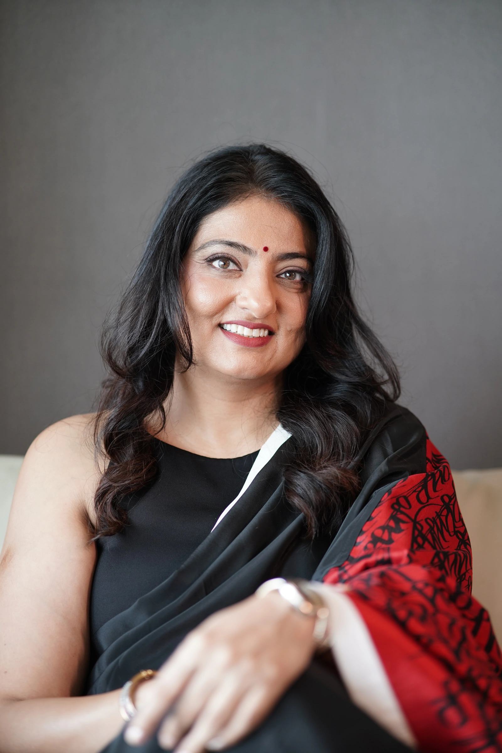 Deval Soparkar smiling in a black sari with a red patterned drape, against the original grey office backdrop.